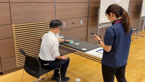 男性社員が机に手を置いて何かを測定し、横で女性が計測している画像