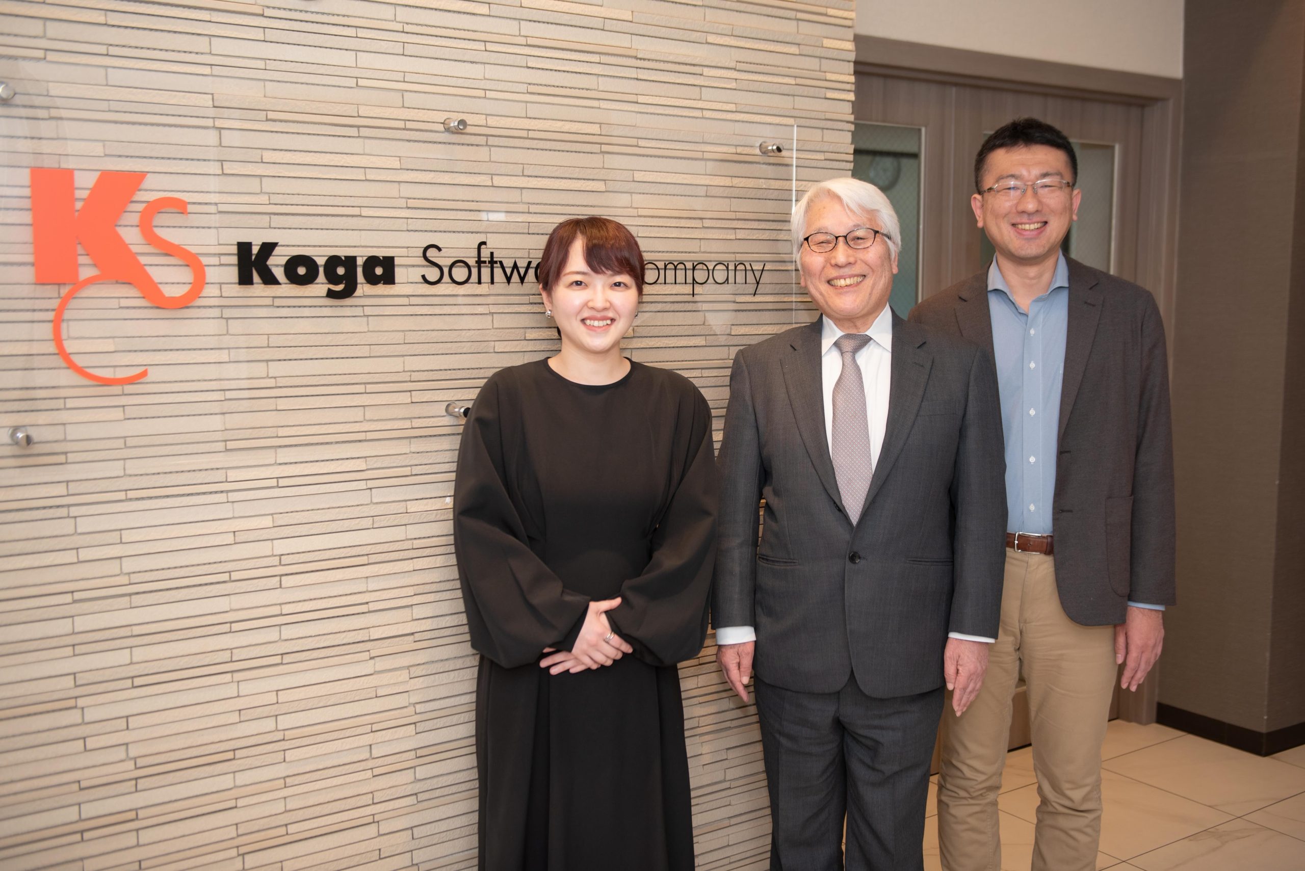 企業看板の前で立っているコガソフトウェア株式会社の箕輪さん、古賀さん、善養寺さん