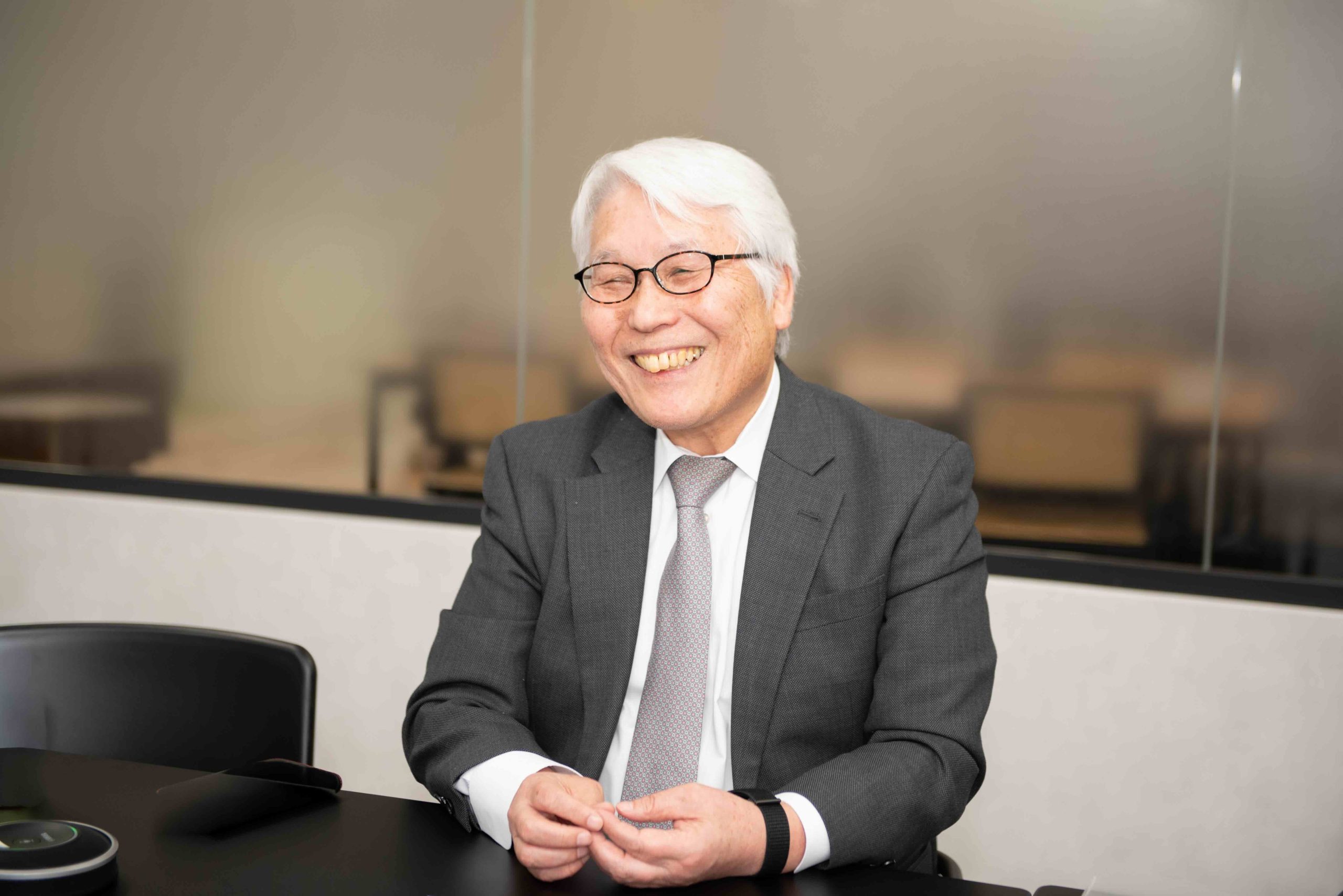 机に手を置いて笑いながらインタビューに応える古賀さんの画像