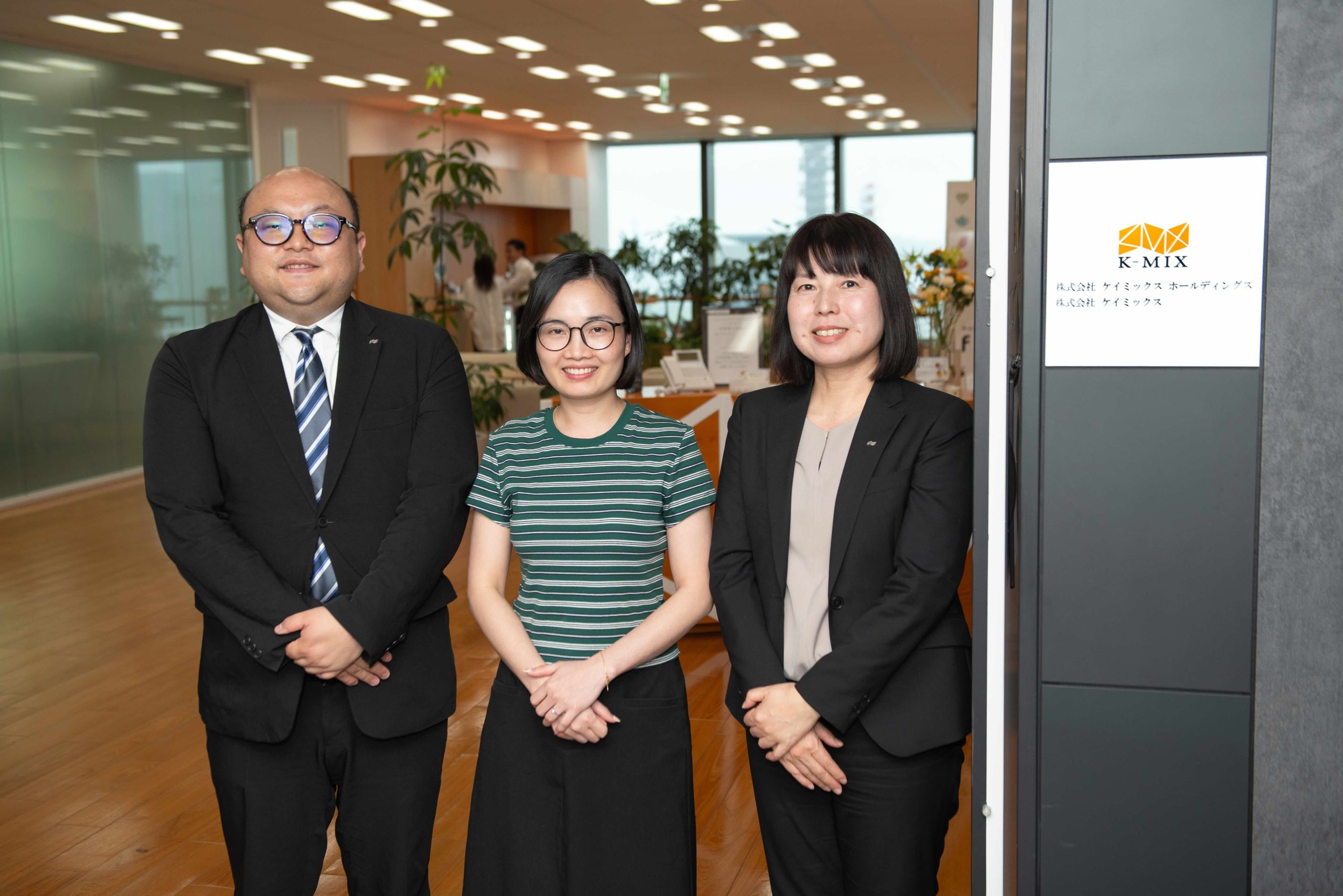 企業看板の前で立っている株式会社ケイミックスの西宮さん、佐藤さん、シンさん